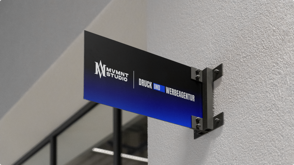 Signboard mounted on a textured wall reading 'MVMT STUDIO' and 'DRUCK UND WERBEAGENTUR' with a blue gradient at the bottom (print and advertising agency).