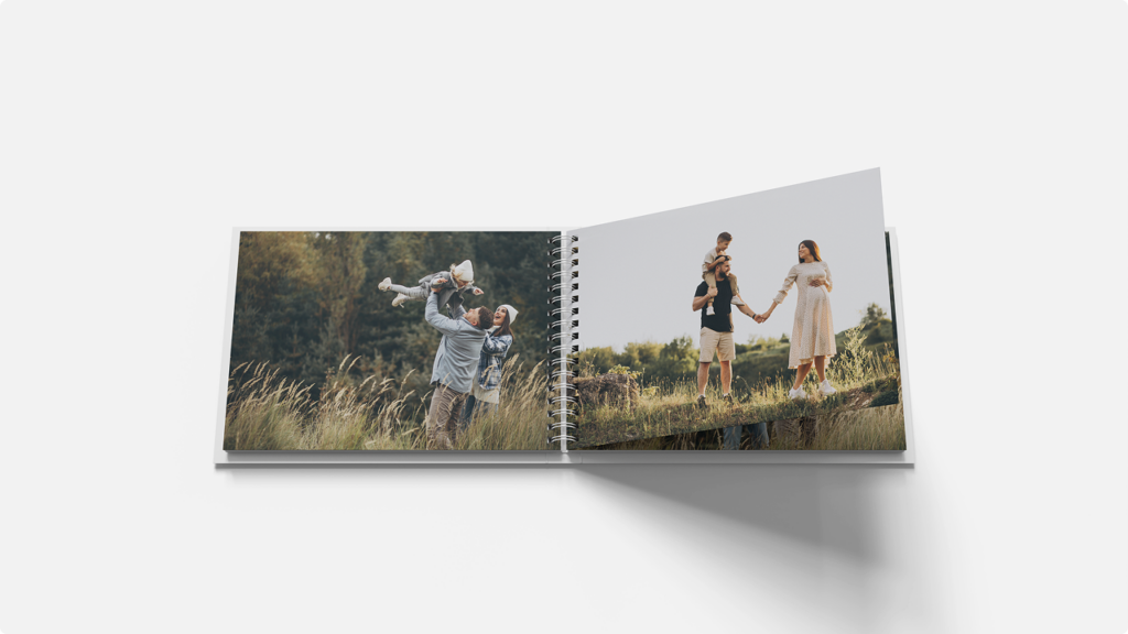 Open photo album on a grassy field; left page shows a man lifting a baby, right page shows a couple holding a child on their shoulders.