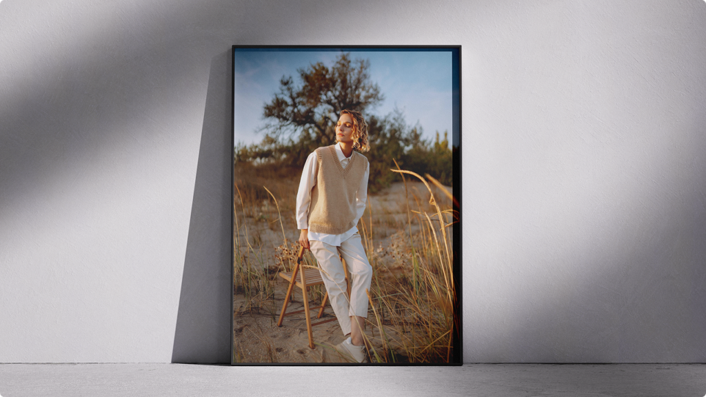 Framed outdoor fashion photo of a woman in a beige knit vest and white outfit standing beside a wooden stool in a sandy field at sunset light