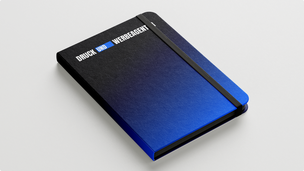 Gradient blue-to-black notebook with a black elastic band laid on a light surface; cover displays the text 'DROCK UND WERBEAGENT'.