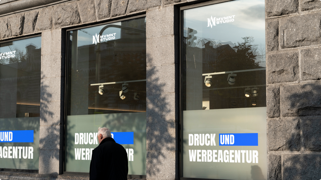 Older man in a black coat walks past a stone storefront with large glass windows showing white text and a blue accent reading 'DRUCK UND WERBEAGENTUR' and a 'MVMNT STUDIO' logo on the glass.